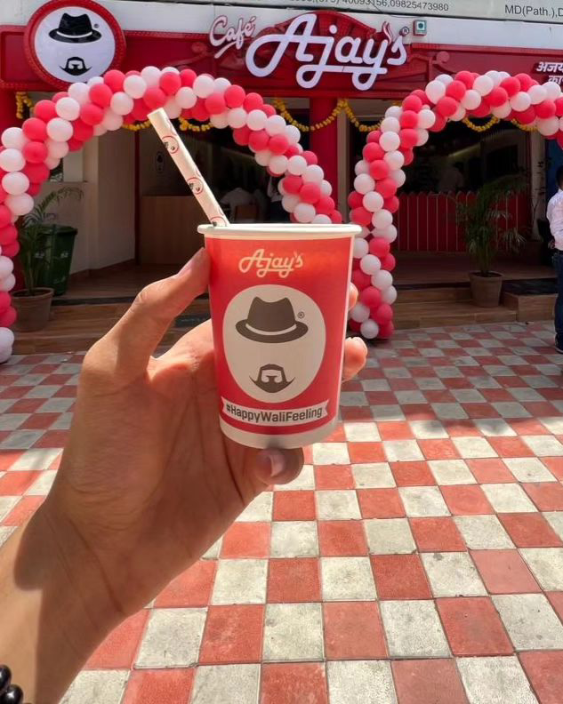 Self-shot image of a person holding Ajay’s cold coffee in a cup that says happy waali feeling. Behind it, Ajay’s cafe is visible.
