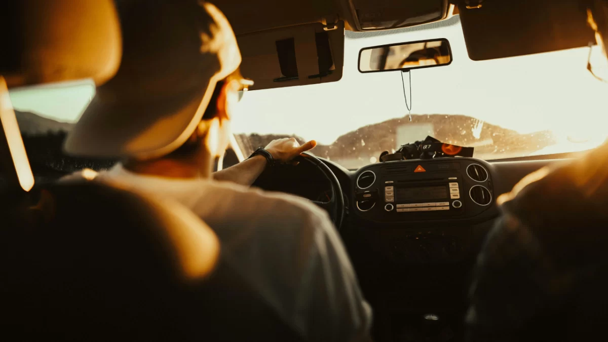 Person driving a car during sunrise with one hand on the steering wheel, sunlight streaming through the windshield on a scenic road.
